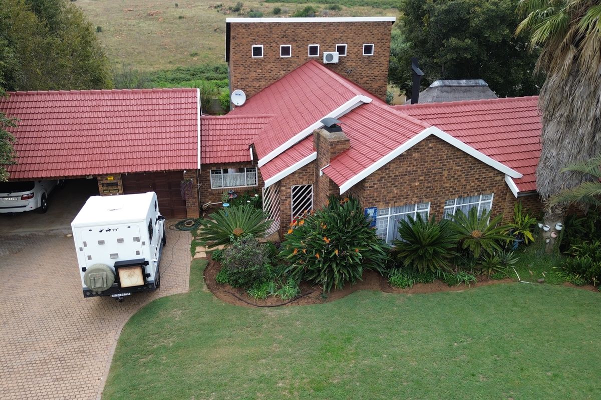 Aerial view of a brick house with a red tiled roof, featuring a paved driveway, garage, and established garden.