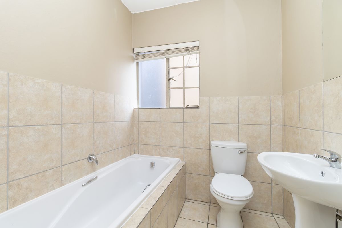 A functional bathroom featuring a built-in bathtub, pedestal basin, and toilet with neutral wall tiling and natural light from a small window.
