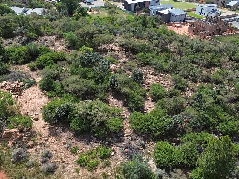 An aerial view of a vacant residential plot featuring natural vegetation and rocky terrain, situated within a developing neighborhood.