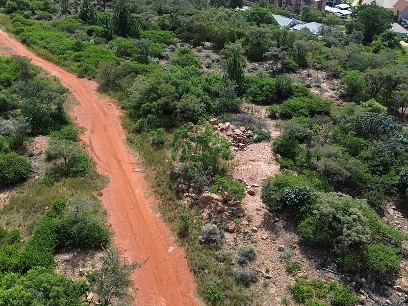 Aerial view of a vacant residential plot featuring natural vegetation, rocky terrain, and an unpaved access road.