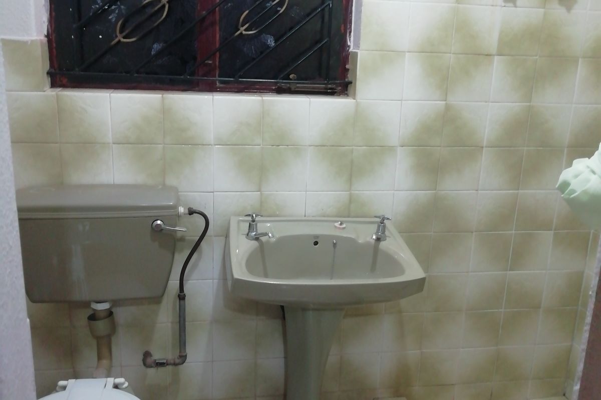 A compact bathroom featuring a pedestal basin, toilet, and shower area with dated wall and floor tiles and a security-barred window.