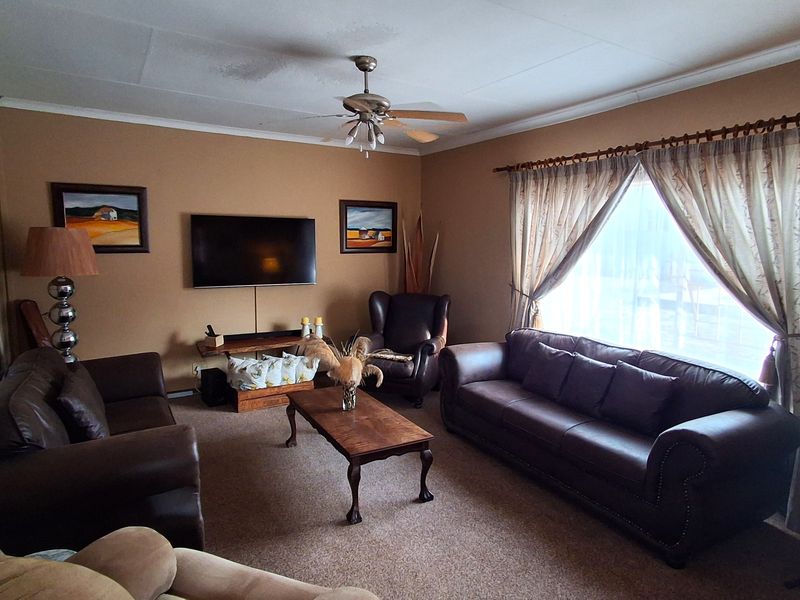 A spacious living room featuring carpeted flooring and a ceiling fan, with visible water staining on the ceiling requiring attention.