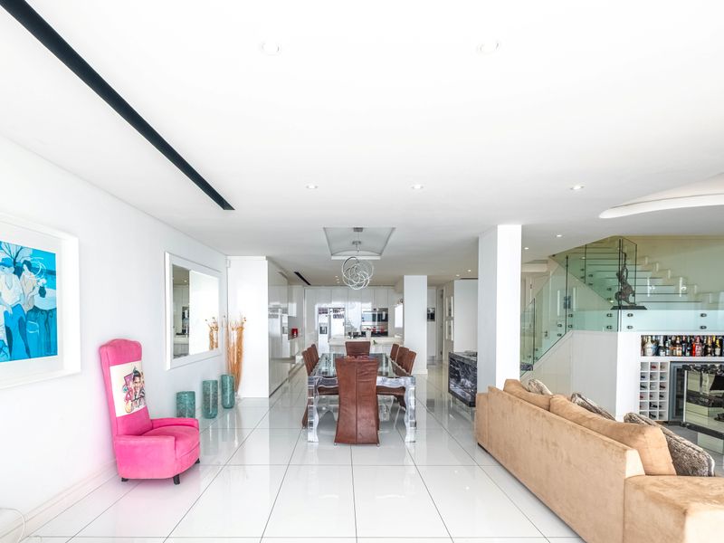Spacious open-plan living and dining area featuring polished white floor tiles, recessed ceiling lighting, and a modern glass staircase with balustrade.