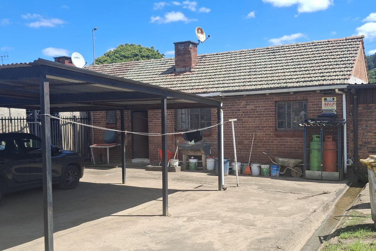 Rear exterior view of a brick house featuring a large carport, concrete driveway, and security fencing, showing signs of wear and weathering.