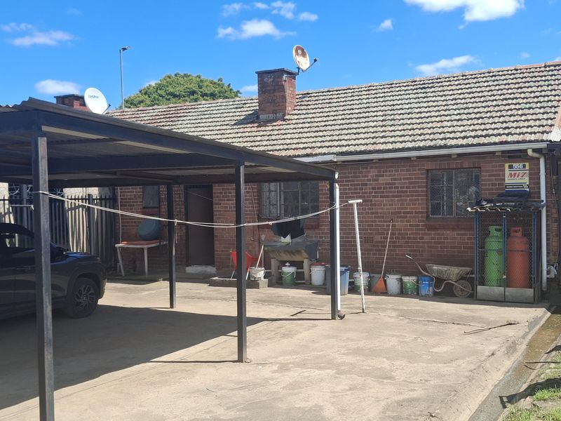 Rear exterior view of a brick house featuring a large carport, concrete driveway, and security fencing, showing signs of wear and weathering.