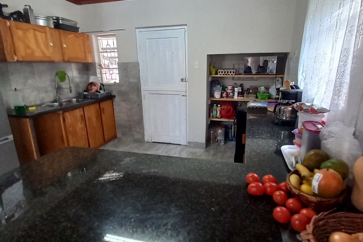 A functional kitchen featuring wooden cabinetry, granite countertops, a stainless steel sink, and built-in pantry shelving with tiled splashbacks.