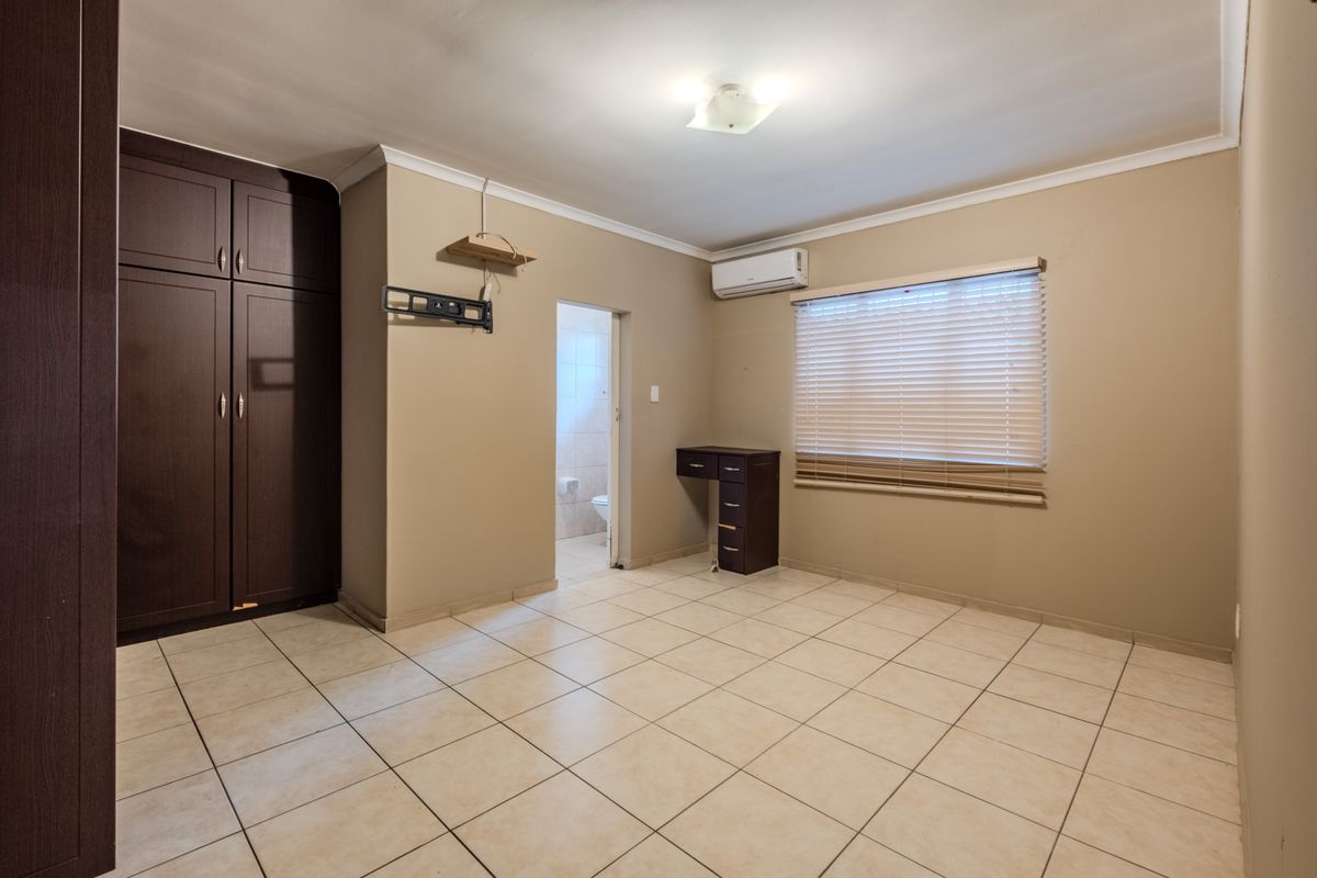 A spacious bedroom featuring neutral tiled flooring, built-in dark wood cupboards, an air conditioning unit, and a window with fitted venetian blinds.