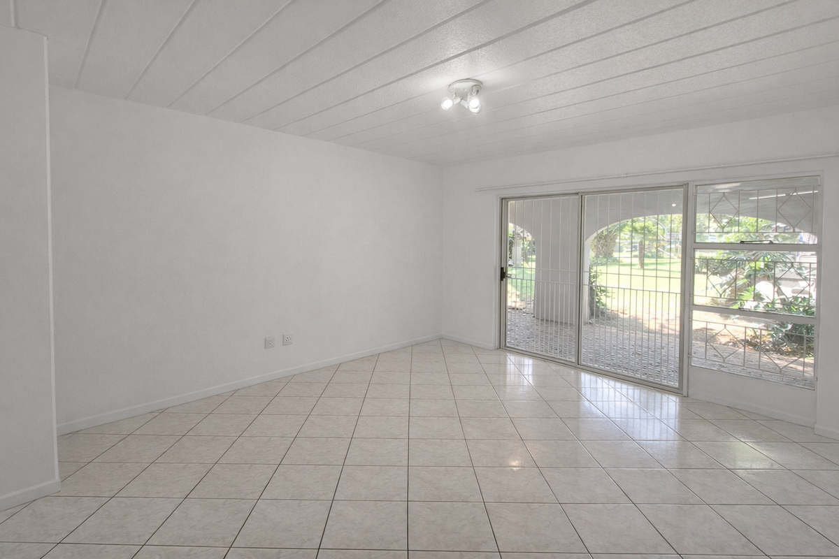 A bright, empty living room featuring tiled flooring, panelled ceilings, and a sliding glass door with security gate leading to the garden.