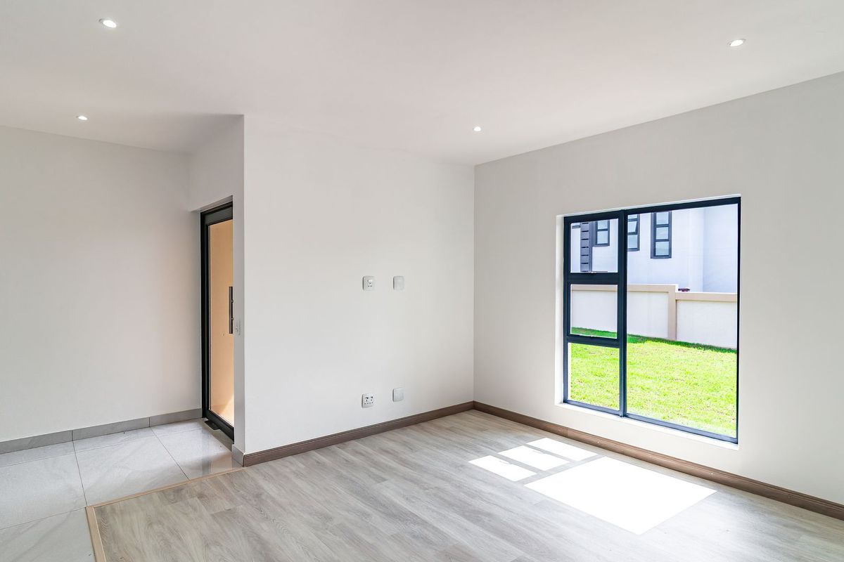 A bright, modern living area featuring neutral wood-look flooring, recessed downlighting, and large aluminum-framed windows overlooking a private garden.