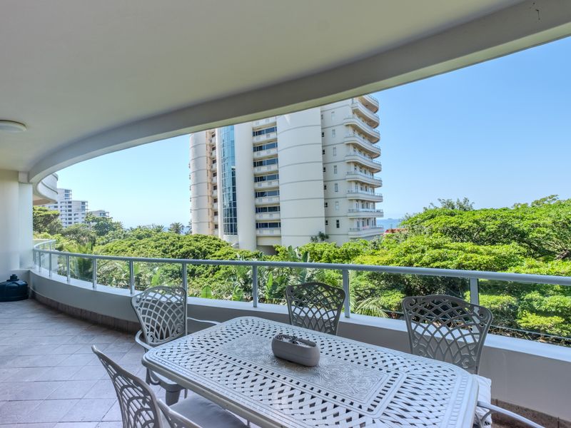 A spacious, curved balcony featuring tiled flooring and a modern glass balustrade, offering pleasant views of the surrounding greenery and nearby buildings.