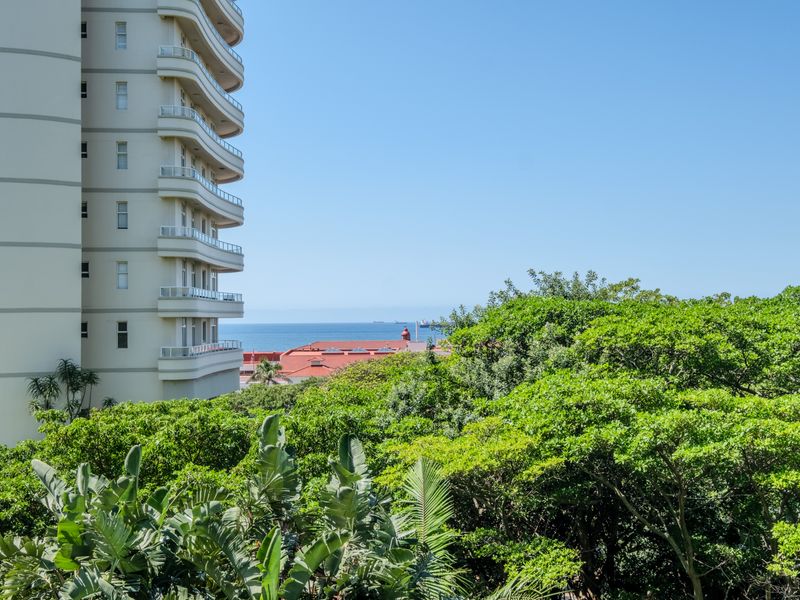 A scenic view overlooking lush green trees with a glimpse of the ocean and a neighbouring high-rise apartment building.