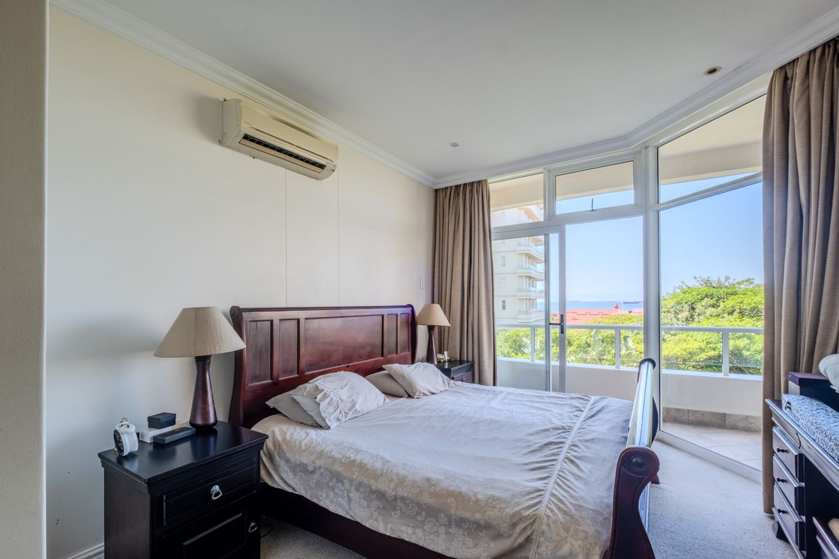A bright bedroom featuring a wall-mounted air conditioning unit, large windows, and sliding glass doors leading to a private balcony with coastal views.