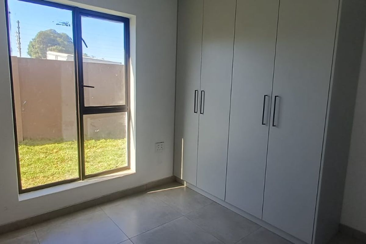 A bright, empty bedroom featuring modern built-in cupboards, neutral tiled flooring, and a large window overlooking the garden area.