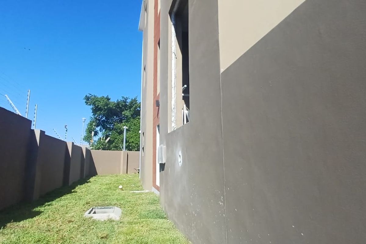 A side view of the apartment building exterior featuring a grassed area, boundary wall with electric fencing, and a manhole cover.