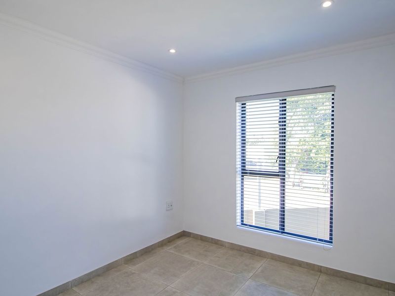 A bright, empty bedroom featuring neutral tiled flooring, recessed downlighting, and a window fitted with venetian blinds.