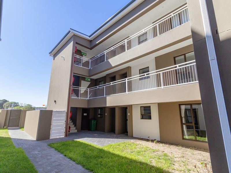 Exterior view of a multi-story apartment building featuring balconies with white railings, paved walkways, and landscaped lawn areas.