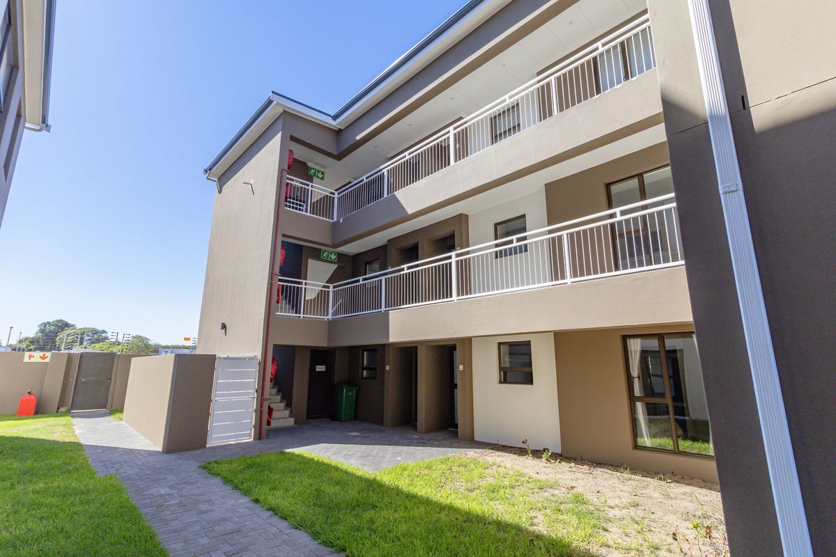 Exterior view of a multi-story apartment building featuring balconies with white railings, paved walkways, and landscaped lawn areas.