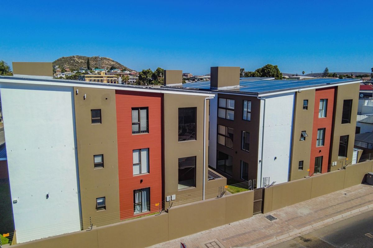 Aerial view of a modern multi-story apartment complex featuring solar panels, perimeter wall with electric fencing, and contemporary exterior finishes.