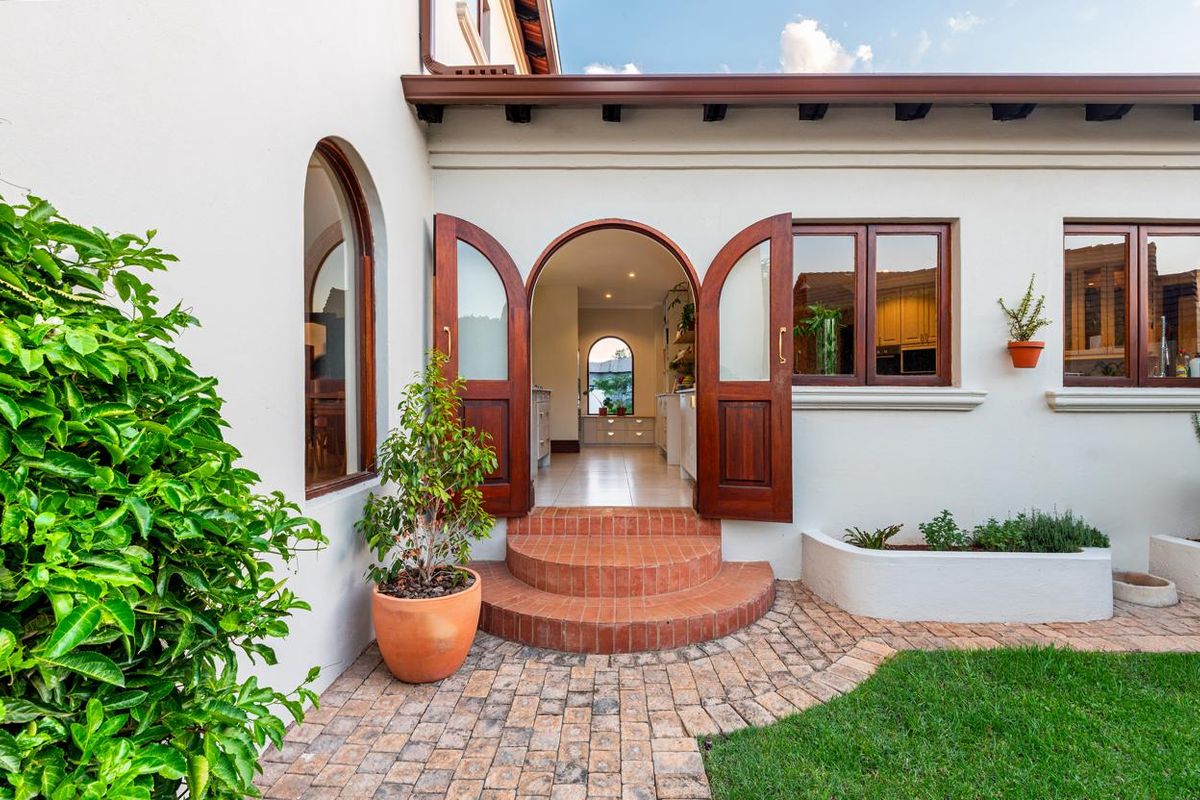 A charming rear exterior view featuring arched wooden double doors, brick steps, and a paved patio area leading into the home.