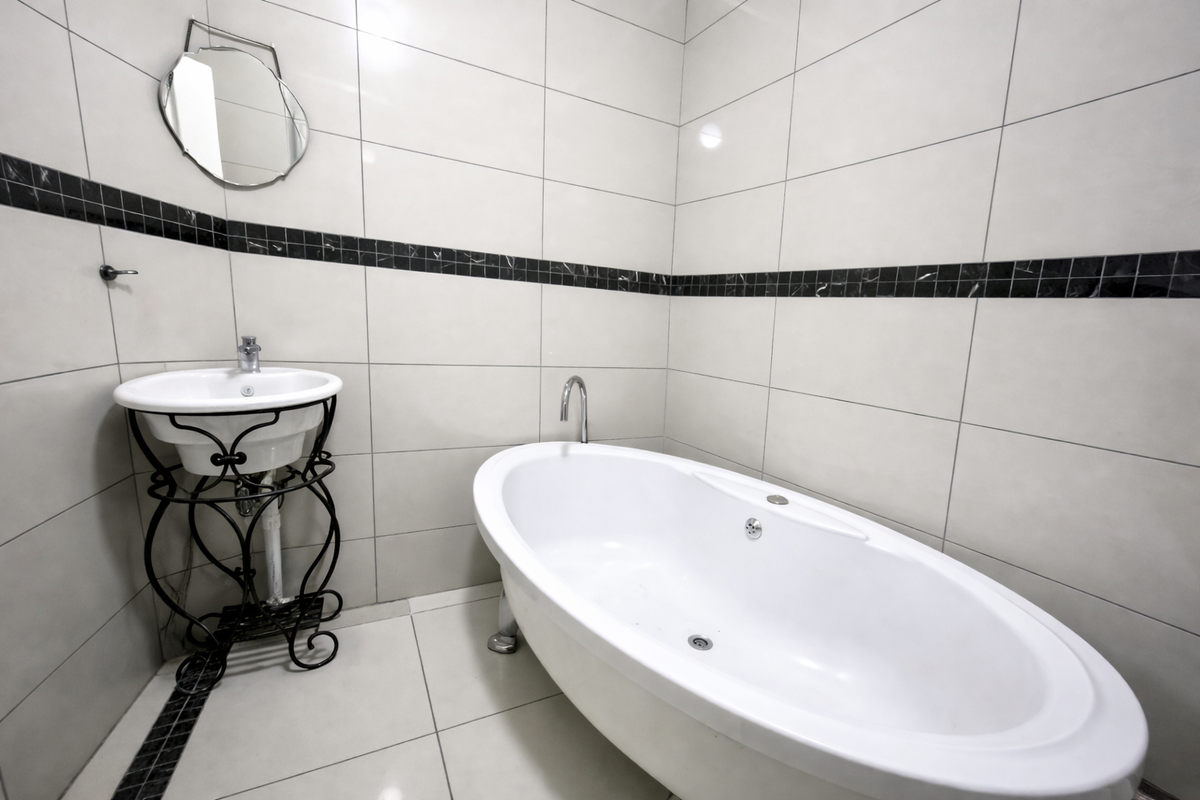 A clean, modern bathroom featuring a white freestanding bathtub, a pedestal washbasin, and neutral wall tiling with a decorative black accent border.
