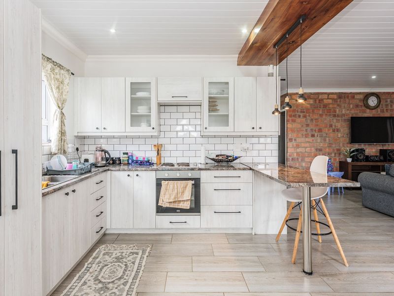Open-plan kitchen featuring white cabinetry, subway tile backsplash, granite countertops, and a breakfast bar, seamlessly flowing into a living area with exposed brick.