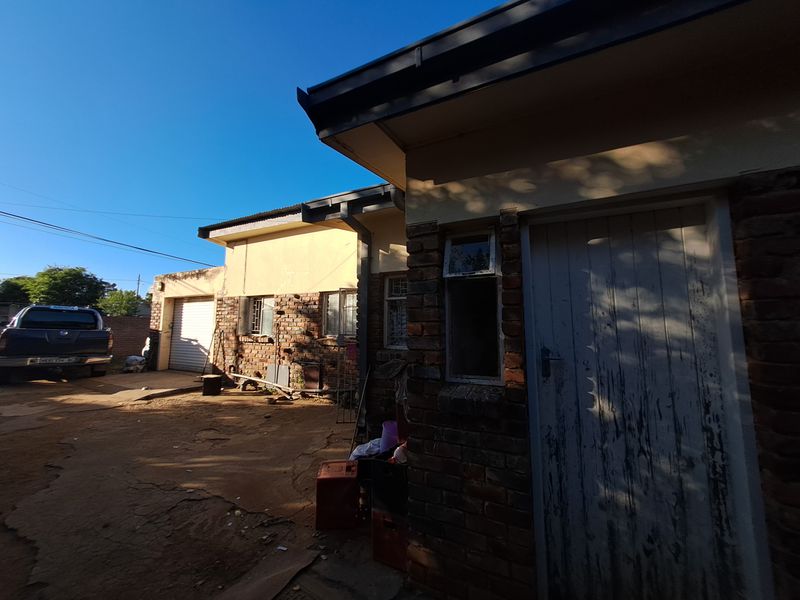 Rear exterior view of the property showing exposed brick walls, a metal garage door, and a weathered timber entrance door with cracked concrete paving.