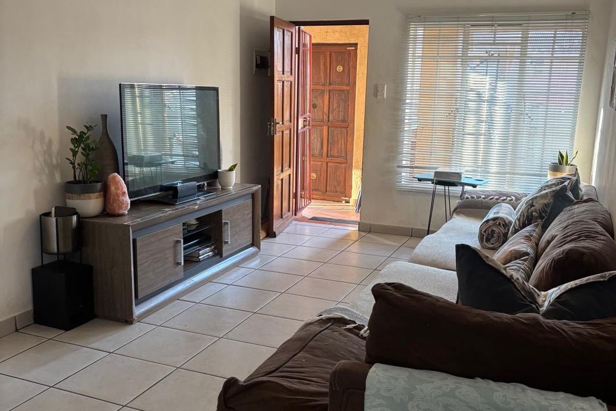 A functional living room featuring ceramic floor tiles, natural light through venetian blinds, and direct access to the exterior via a security gate.