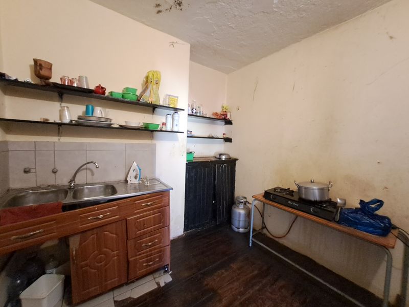 A dated kitchen area featuring basic wooden cabinetry, open shelving, and a stainless steel sink, requiring significant renovation and cosmetic updates.