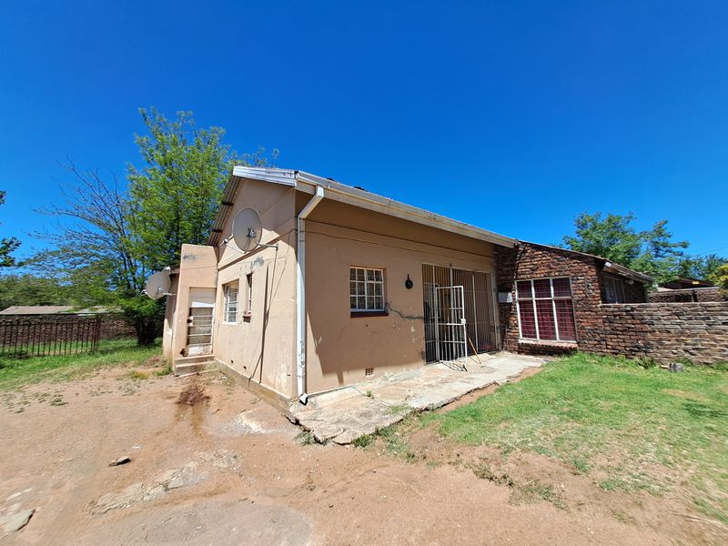 Rear exterior view of a single-story house featuring brick and plastered walls, security gates, and a concrete patio area requiring maintenance.
