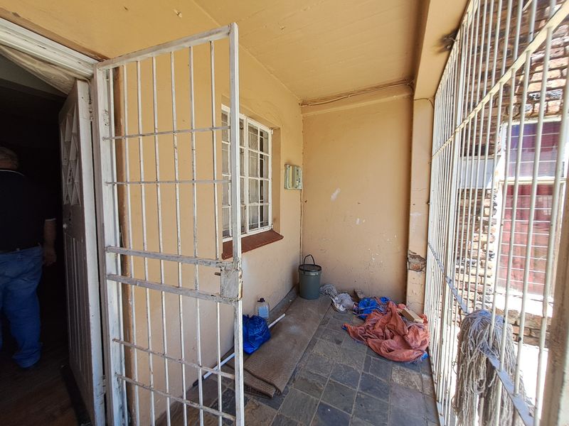 A covered patio area featuring tiled flooring, security gates, and protective bars, requiring some maintenance to the wall finishes.