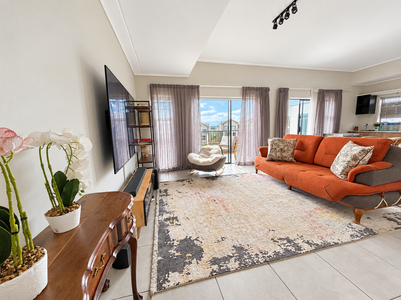 A bright and spacious living room featuring large sliding glass doors that open to a balcony, complemented by modern track lighting and tiled floors.