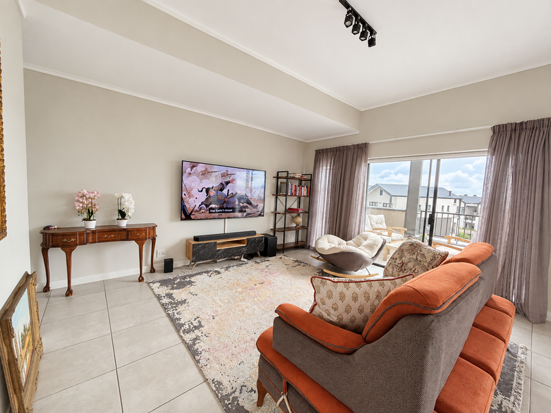 A bright and spacious living room featuring tiled flooring, modern track lighting, and large sliding glass doors that open onto a balcony.