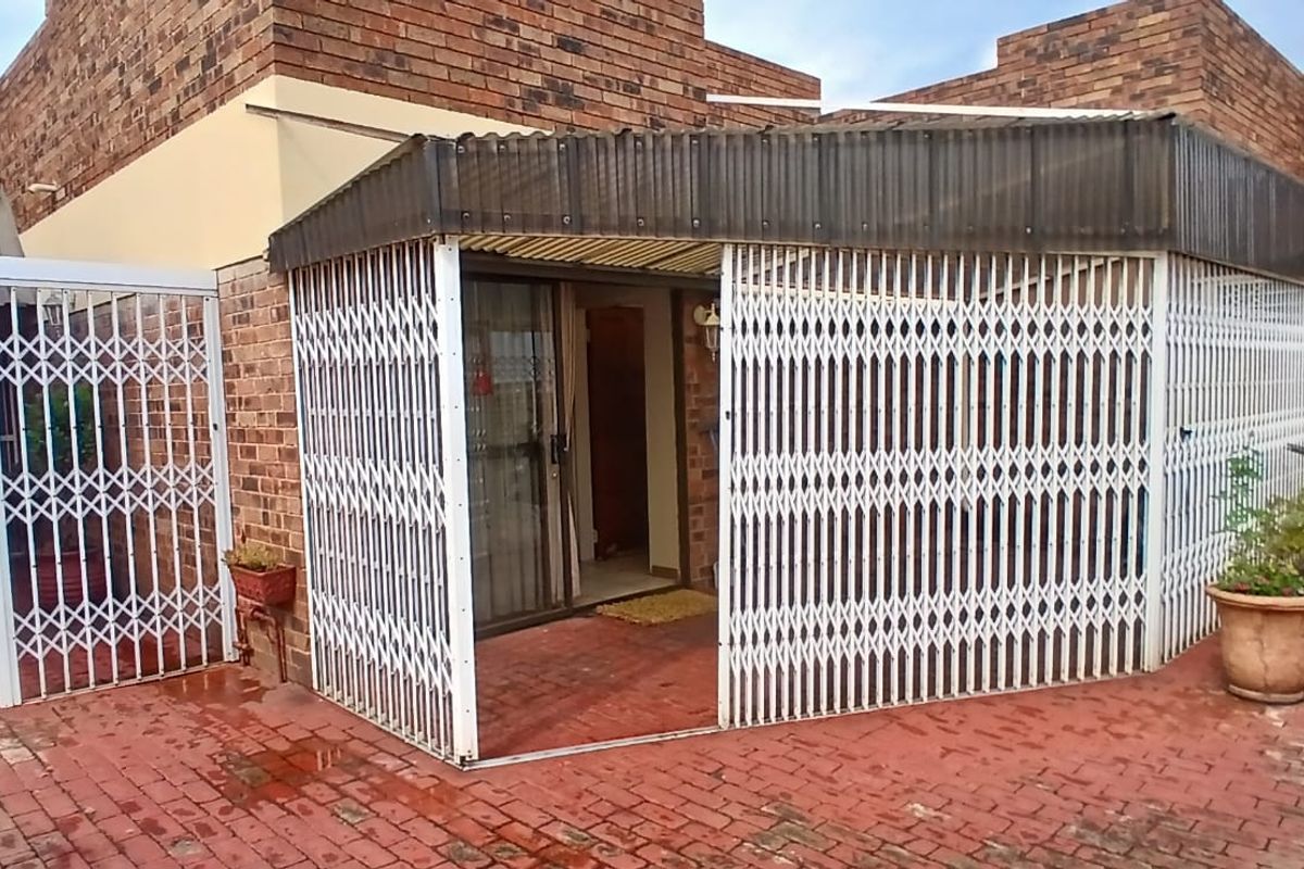 A rear exterior view of the flat featuring a paved courtyard area enclosed by white metal security gates and a covered patio structure.