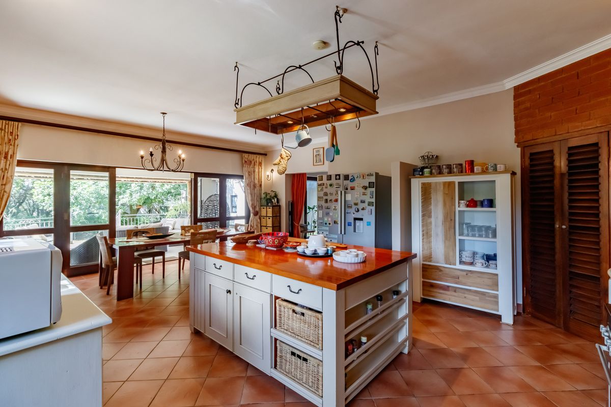 Spacious kitchen featuring a large central island with a wooden countertop, hanging pot rack, and terracotta flooring, flowing into an adjacent dining area.