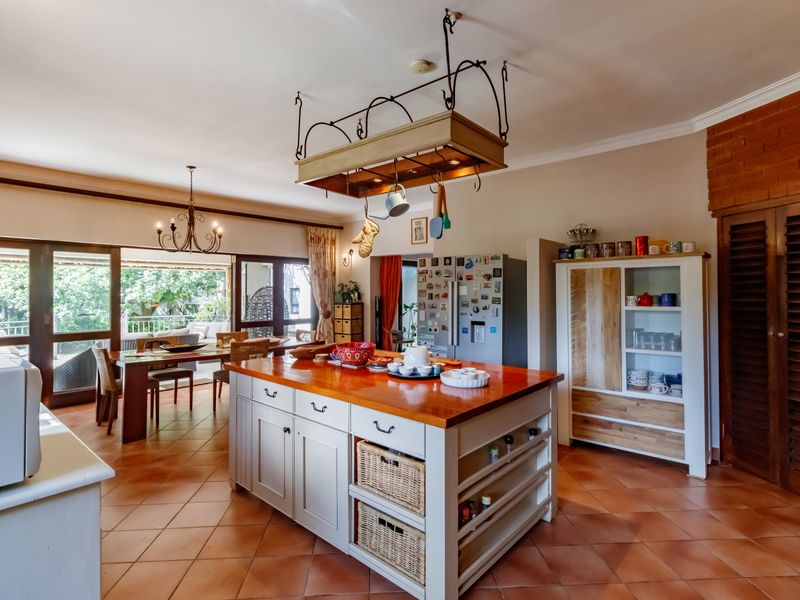 Spacious kitchen featuring a large central island with a wooden countertop, hanging pot rack, and terracotta flooring, flowing into an adjacent dining area.