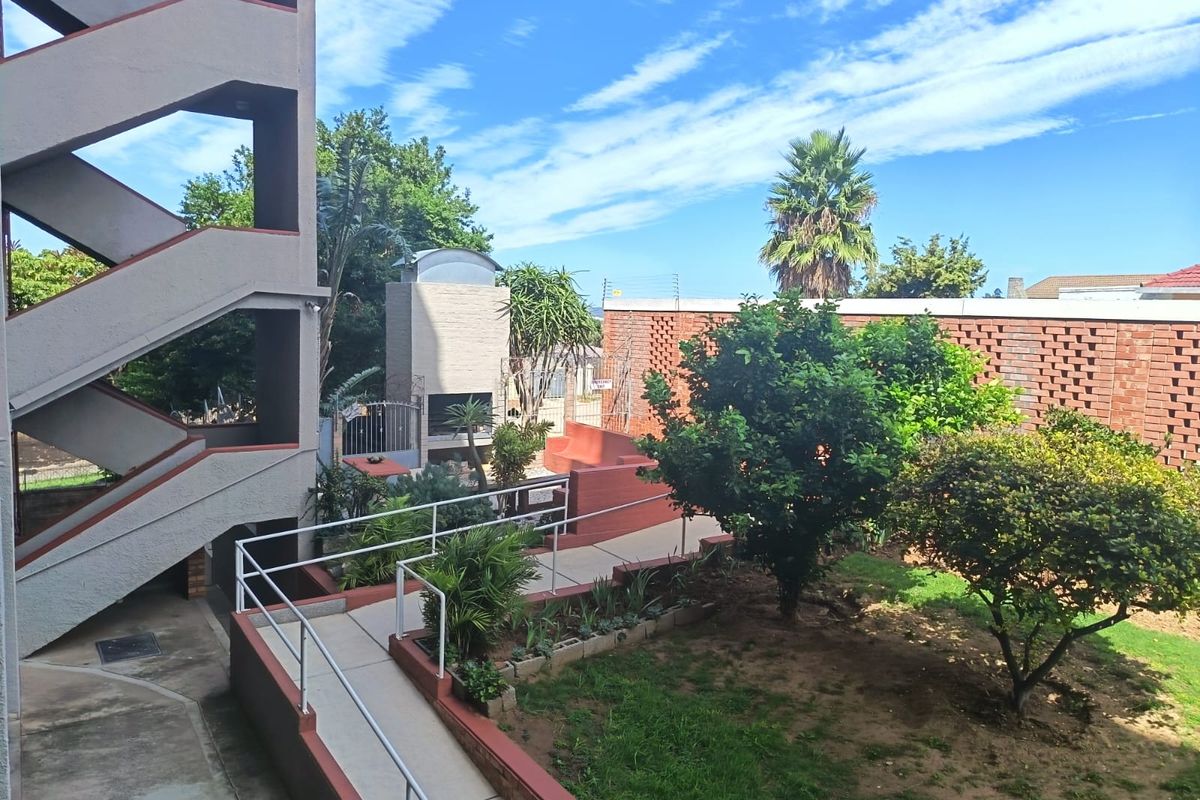 A communal outdoor area featuring a built-in braai, paved walkways with metal handrails, and landscaped garden space within a secure brick-walled complex.