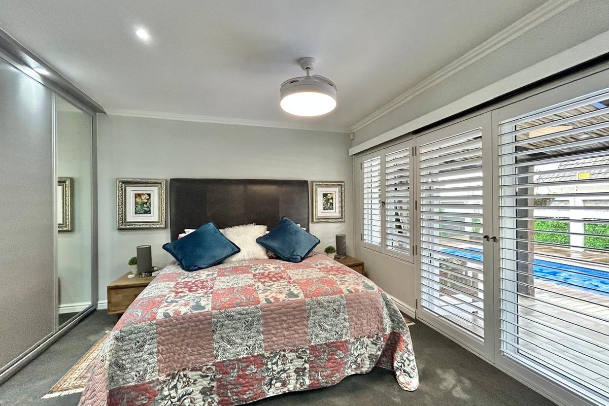 A well-maintained bedroom featuring built-in mirrored wardrobes, a modern ceiling fan, and plantation shutters opening to an outdoor pool area.