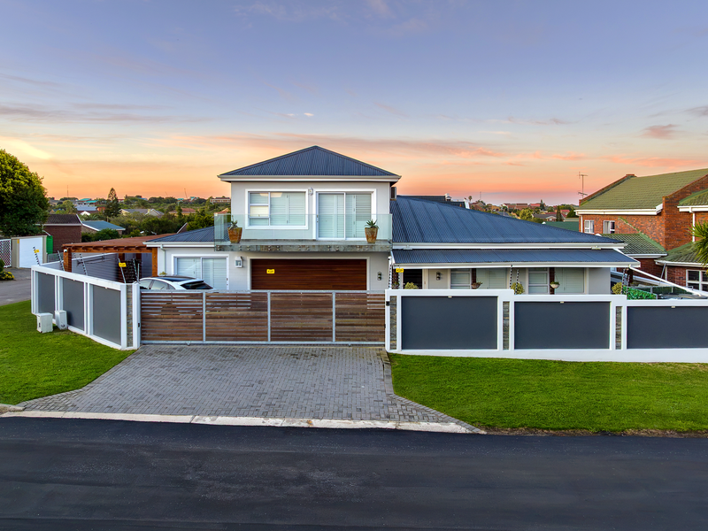 Modern double-story residential property featuring a paved driveway, secure perimeter wall with electric fencing, timber garage door, and an upper-level balcony.