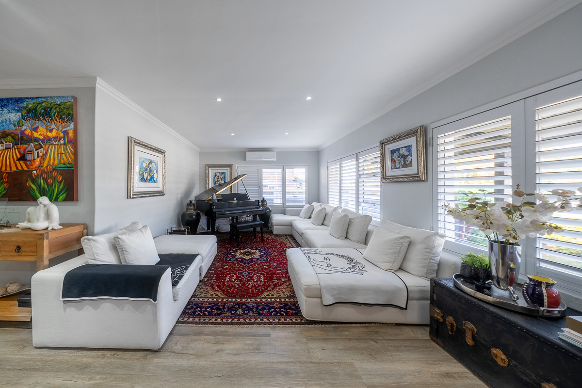 A bright and spacious living room featuring laminate flooring, recessed downlighting, a wall-mounted air conditioning unit, and plantation shutters.
