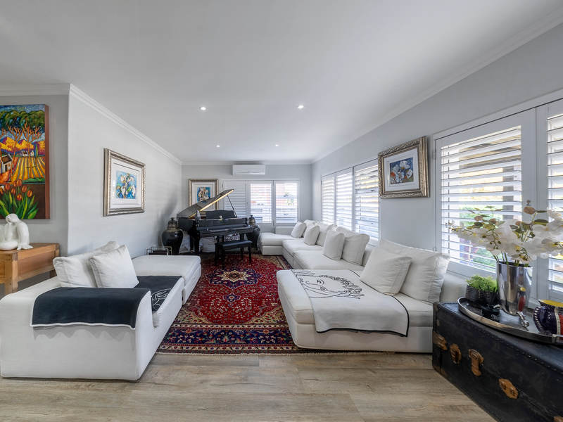 A bright and spacious living room featuring laminate flooring, recessed downlighting, a wall-mounted air conditioning unit, and plantation shutters.