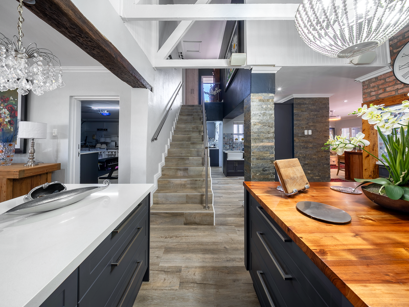 A modern open-plan kitchen and living area featuring exposed timber beams, stone-clad pillars, a central staircase, and a mix of quartz and timber countertops.