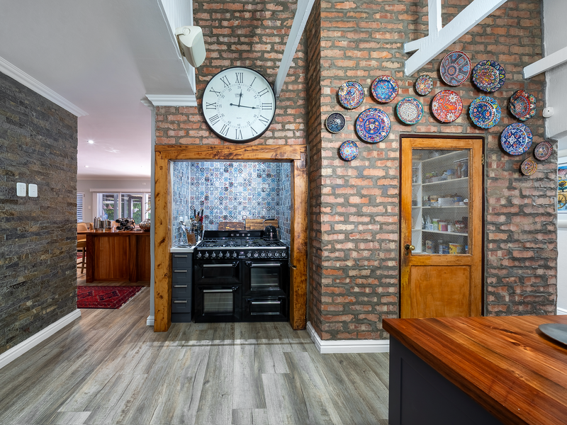 A characterful kitchen featuring an exposed brick wall, a built-in range cooker with decorative tiled splashback, and a dedicated pantry with a timber door.