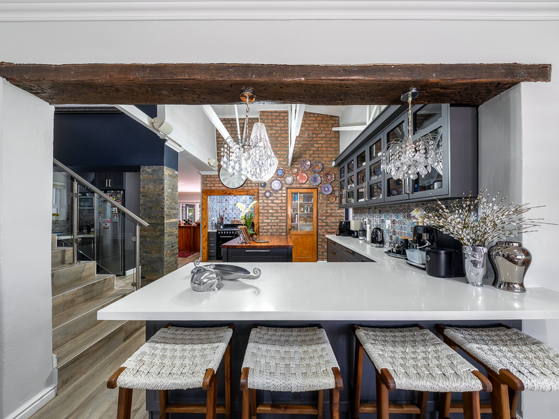 Modern kitchen featuring a large quartz breakfast bar, exposed brick feature wall, and elegant glass-fronted cabinetry, seamlessly connected to an open-plan living area.