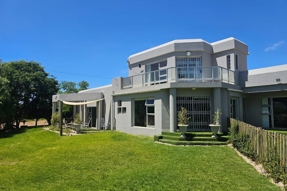 Rear view of a multi-level farm residence featuring a spacious lawn, covered patio area, and an upper-level balcony with glass balustrades.