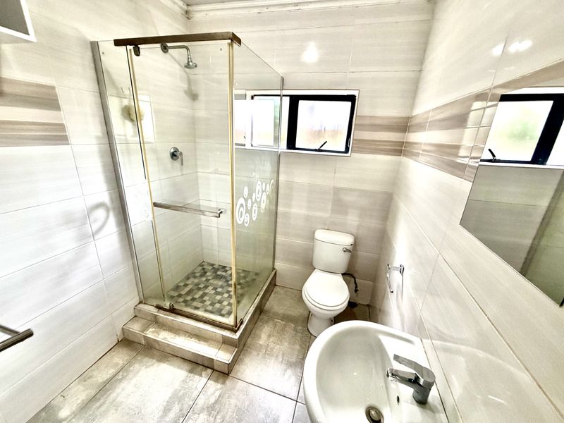 A functional bathroom featuring a glass-enclosed shower, pedestal basin, and toilet with neutral wall and floor tiling.