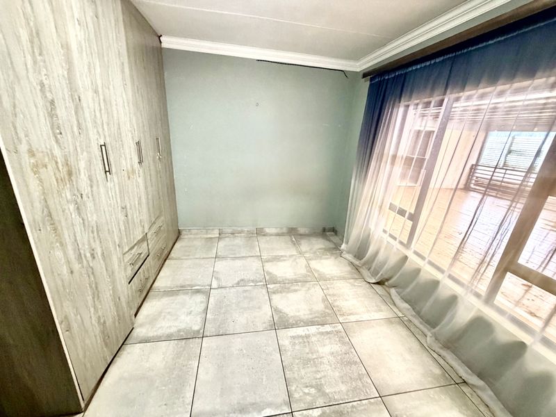 A bright, empty bedroom featuring modern wood-look built-in cupboards, neutral tiled flooring, and a large sliding door providing natural light.