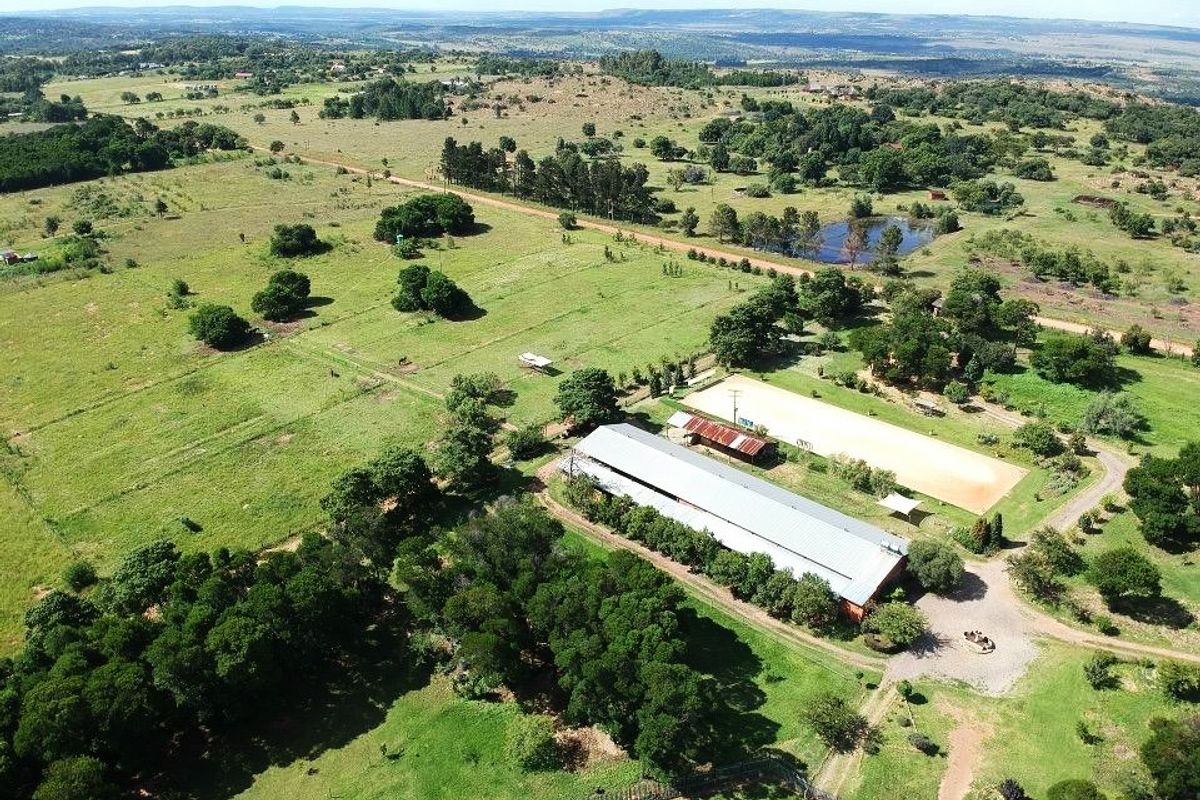 Aerial view of a small holding featuring a large agricultural shed, an equestrian arena, a farm dam, and extensive open grazing land.