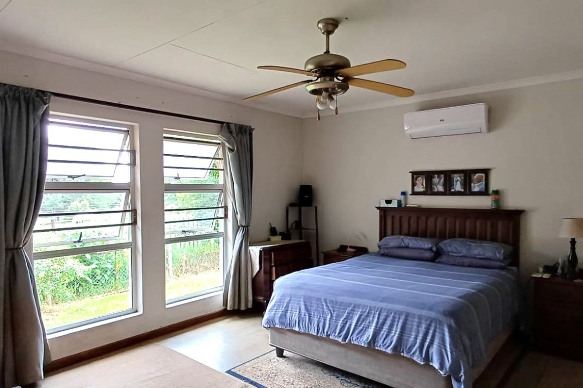 A spacious bedroom featuring a ceiling fan, wall-mounted air conditioning unit, and large louver windows providing natural light.