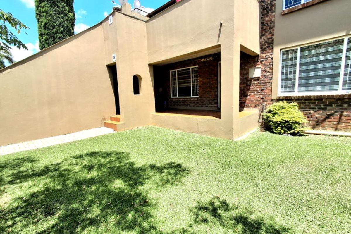 A rear view of the townhouse featuring a covered patio area, brick exterior walls, and a well-maintained private lawn.