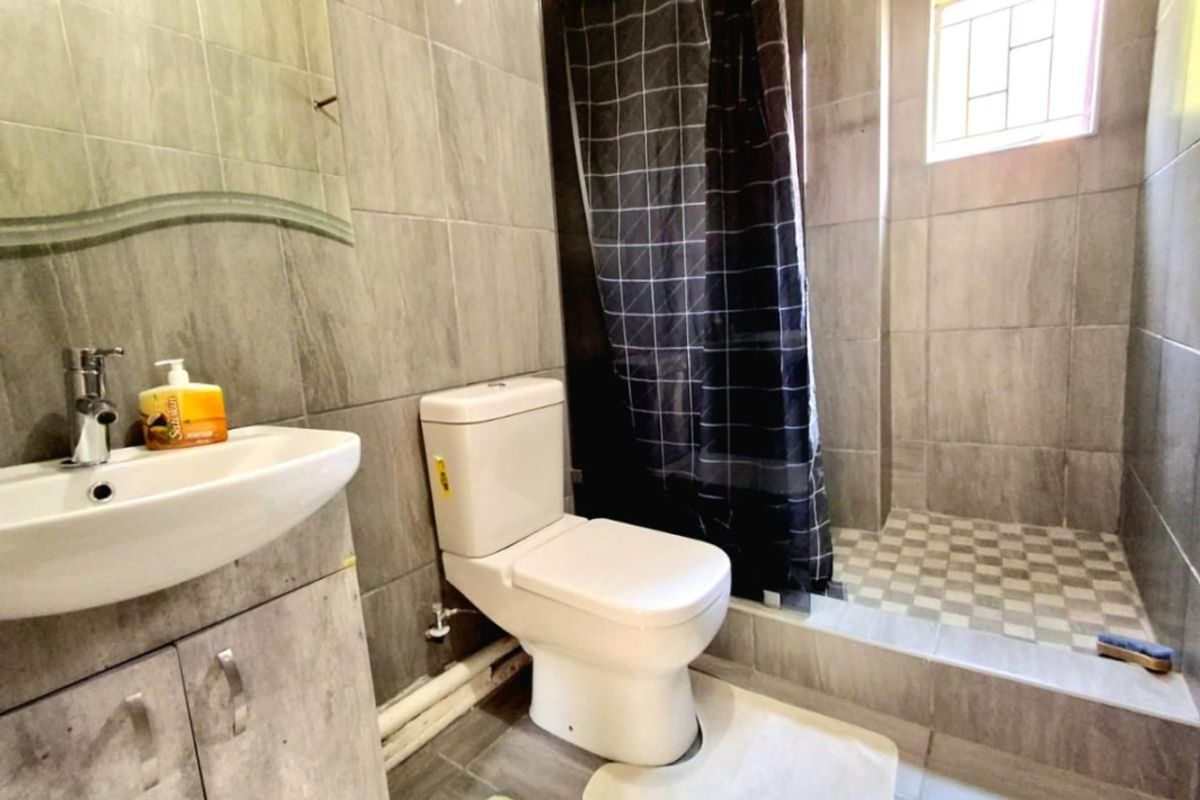 A functional bathroom featuring grey wall tiles, a vanity basin, a toilet, and a walk-in shower area with a privacy curtain.